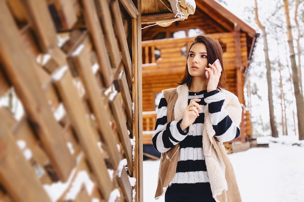 Chica joven con teléfono en chaleco en contra de casa de madera en bosque de invierno