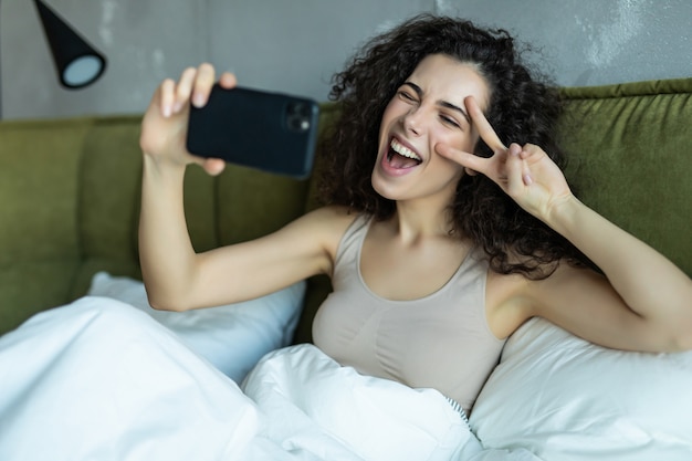 Chica joven tomando selfie desde teléfono con gesto de paz por la mañana en la cama