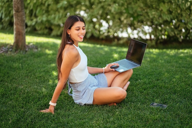 La chica joven trabaja con una computadora portátil sentada en el césped