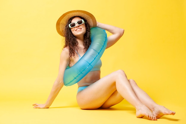 Chica joven en traje de baño azul y sombrero de paja con un círculo de natación inflable se sienta en la playa