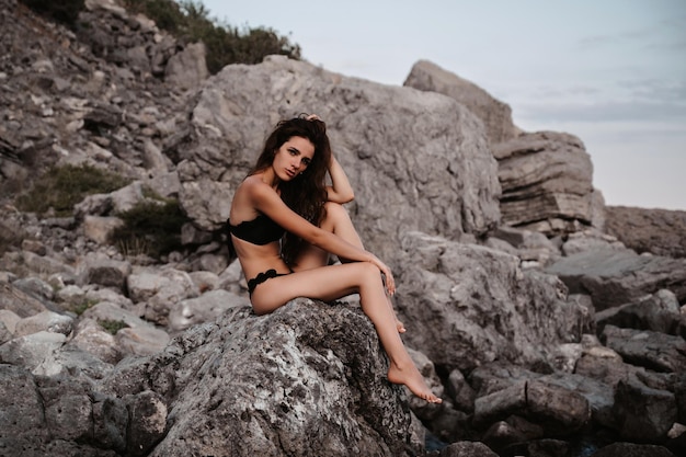 Chica joven en traje de baño negro está sentada sexy en las rocas en la playa junto al mar en verano