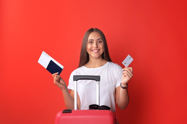 Chica joven turista en ropa casual de verano, con gafas de sol, maleta roja, pasaporte aislado sobre fondo rojo.