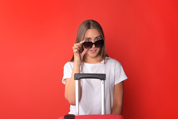 Chica joven turista en ropa casual de verano, con gafas de sol, maleta roja, pasaporte aislado sobre fondo rojo.