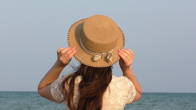 Chica joven con vestido de encaje blanco y sombrero de pie en la playa tropical. Mar azul en el fondo. Concepto de vacaciones de verano