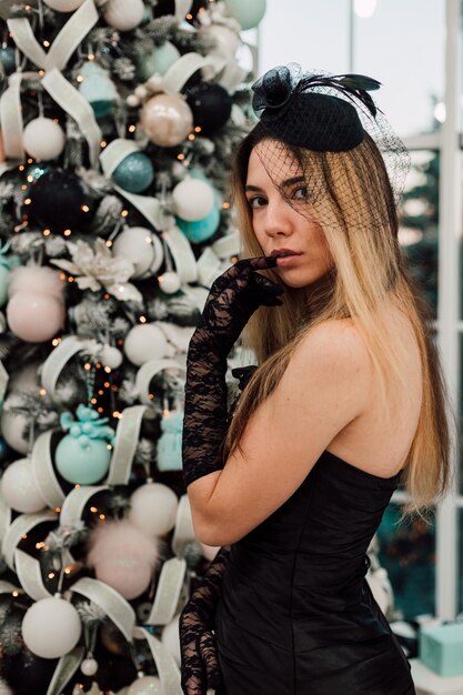 Chica joven con un vestido largo negro posando cerca de un árbol de Navidad decorado