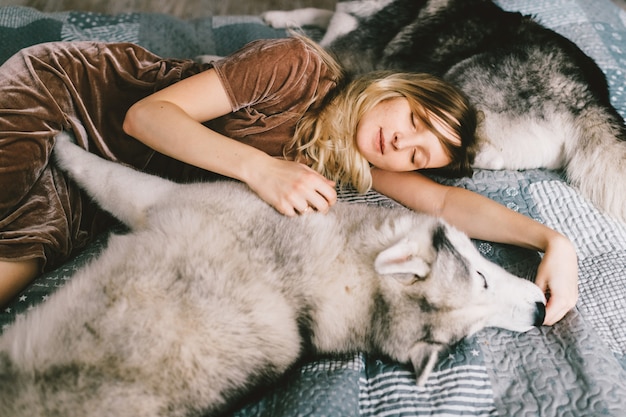 Chica joven en vestido marrón acostado en cama en casa y durmiendo con cachorro husky. El retrato interior de la forma de vida de la mujer hermosa abraza el perro fornido en el sofá. Amante de mascotas. Alegre mujer descansando con adorables perros.