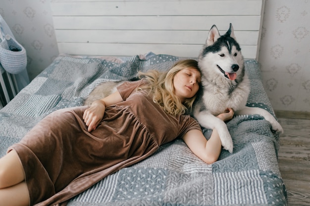 Chica joven en vestido marrón acostado en cama en casa y durmiendo con cachorro husky.