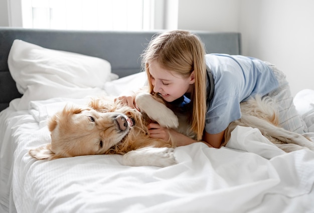 La chica juega con el golden retriever en la cama por la mañana en una habitación luminosa