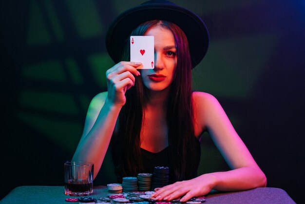 Chica de juego con un sombrero juega al póquer en una mesa con cartas con luces rojas y fondo oscuro