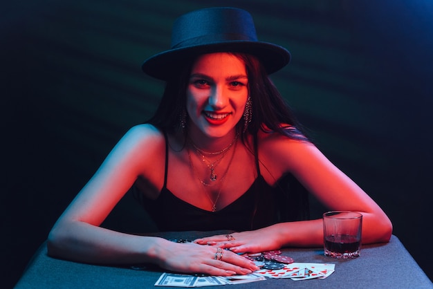 Chica de juego con un sombrero juega al póquer en una mesa con cartas con luces rojas y fondo oscuro