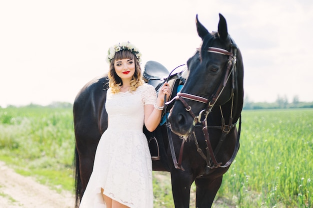 Chica con labios rojos en un vestido blanco cerca de un caballo negro