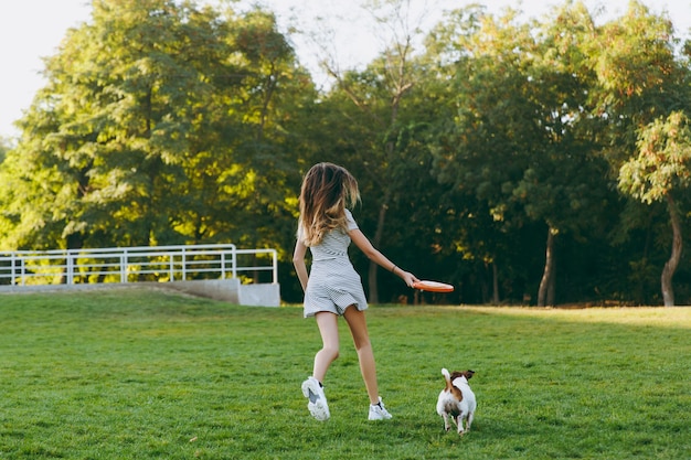 Chica lanzando un disco volador naranja a un pequeño perro gracioso, que lo atrapa en la hierba verde. Pequeña mascota Jack Russel Terrier jugando al aire libre en el parque. Perro y dueño al aire libre.