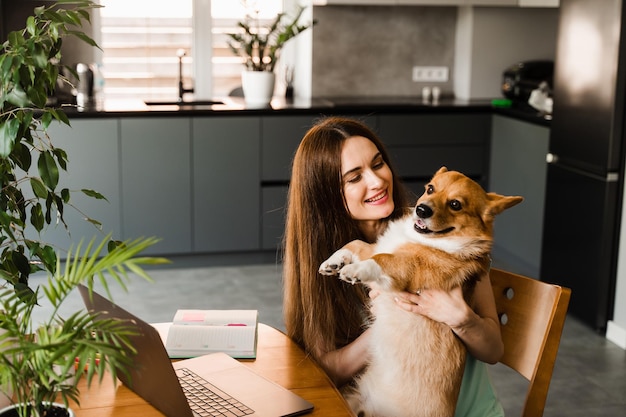 Chica con laptop sonríe y juega con el perro Corgi en casa Programadora que trabaja en línea y toma un descanso para abrazar a su perro Divirtiéndose con Welsh Corgi Pembroke