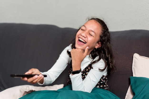 Chica latina divertida riendo y viendo la televisión en el sofá con control remoto en la mano.