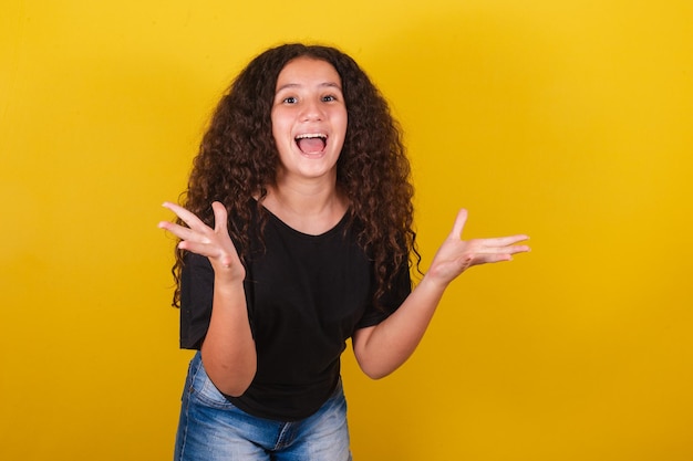 Chica latinoamericana brasileña para cabello afro fondo amarillo sonriendo haciendo expresión sorprendida con cara y manos wow sorprendido