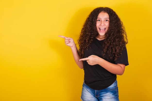 Chica latinoamericana brasileña para cabello afro fondo amarillo sonriendo sugiriendo indicación de sugerencia apuntando con las manos al lado espacio negativo para publicidad y texto publicitario recomendando
