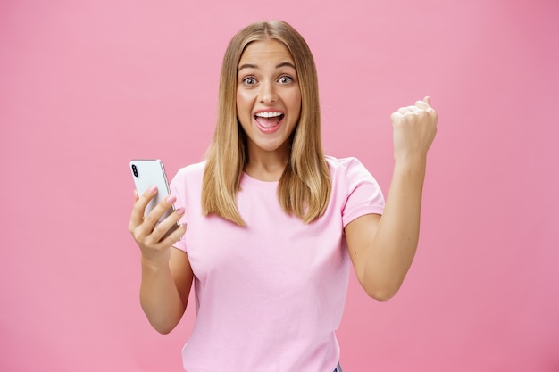 Chica levantando el puño cerrado en alegría y triunfo sosteniendo el teléfono celular, sonriendo emocionado y feliz a la cámara celebrando con gesto alegre sobre fondo rosa