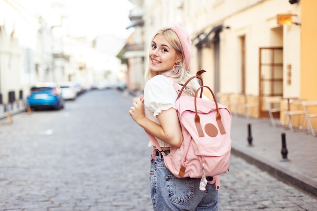 Una chica linda con boina y mochila mira a la cámara y sonríe en una calle europea