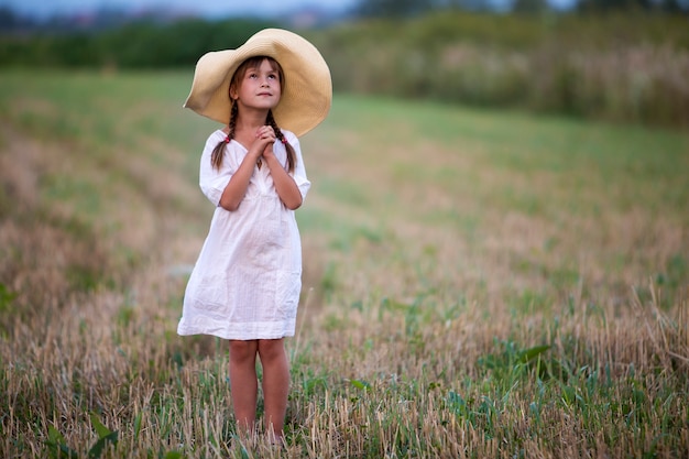 Chica linda encantadora joven de moda con largas trenzas en un bonito vestido blanco de verano y un gran sombrero de paja.