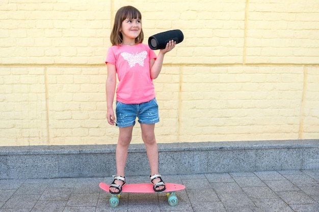 Una chica linda escucha música y monta una patineta contra un fondo de paredes de ladrillo amarillo