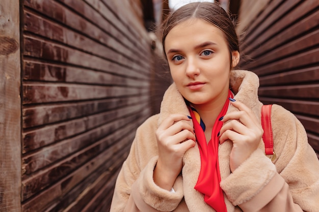 Chica linda con estilo joven en un abrigo de piel paseando por la ciudad cerca de casas de madera y paredes de piedra