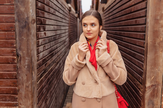 Chica linda con estilo joven en un abrigo de piel paseando por la ciudad cerca de casas de madera y paredes de piedra