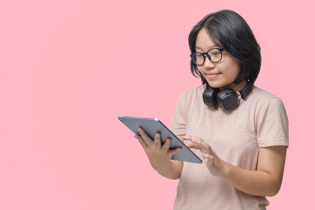 Chica linda con gafas jugando a la tableta con auriculares aislados sobre un fondo rosa