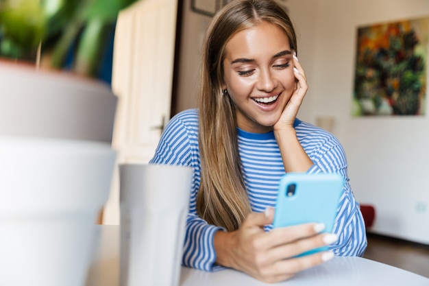 una chica linda joven sonriente optimista alegre en casa en la mesa usando el teléfono móvil.