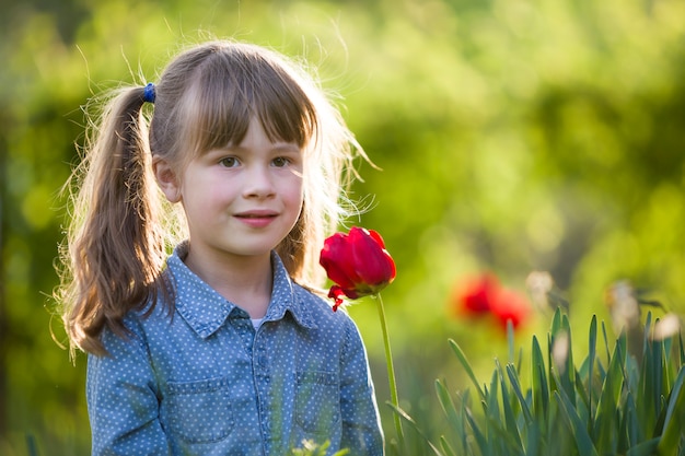 Chica linda niña muy sonriente con ojos grises y cabello largo con flor de tulipán rojo brillante en bokeh verde soleado borrosa. Concepto de naturaleza.