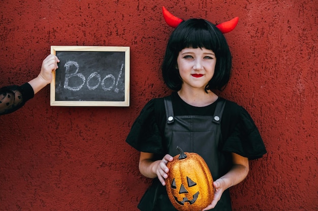 Chica linda con una peluca con cuernos rojos sosteniendo una calabaza sobre un fondo de pared roja Halloween ce