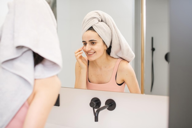 Una chica linda con una toalla en la cabeza se mira en el espejo del baño después de un cuidado de la piel de la ducha