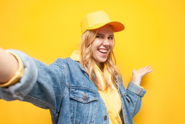 Chica loca con chaqueta de mezclilla y gorra hace selfie sobre fondo amarillo aislado