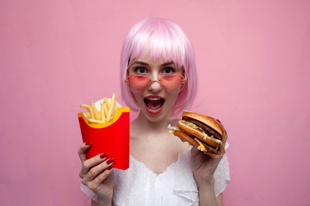 Una chica loca con el pelo corto rosa sostiene una hamburguesa y patatas fritas y grita con un fondo rosa