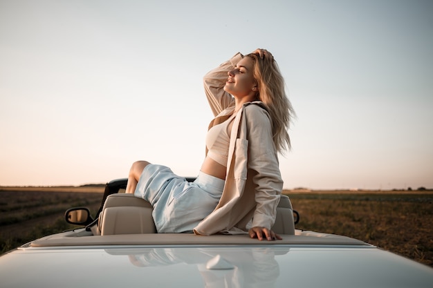 Una chica de lujo glamorosa con cabello rubio sonriendo mientras está sentada en un convertible blanco. Mujer de éxito joven sentada en su coche blanco