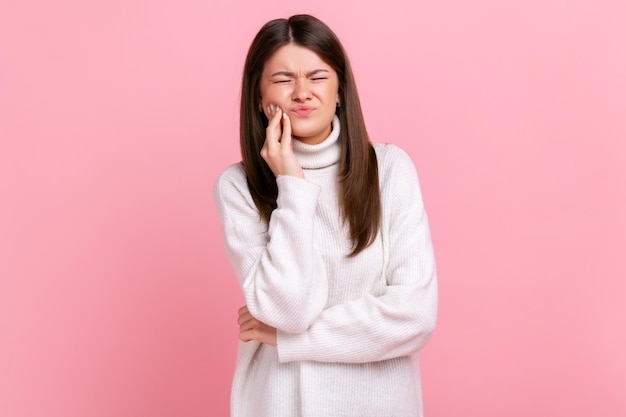 Chica malsana tocando la mejilla, cerrando los ojos con expresión de terrible sufrimiento por problemas dentales, usando suéter blanco de estilo casual. Disparo de estudio interior aislado sobre fondo rosa.