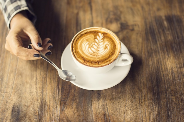 Chica mano manteniendo una cucharadita y una taza de capuchino en una mesa de madera