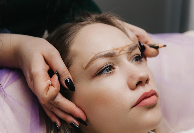 Chica con maquillaje permanente en las cejas La maquilladora hace marcas con un lápiz marrón para tatuarse las cejas