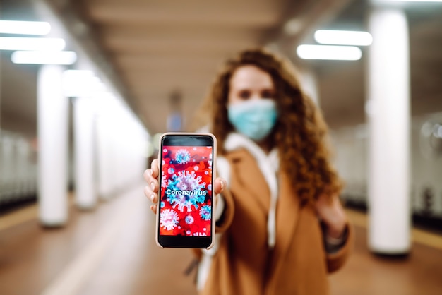 Foto chica con máscara médica protectora estéril en su rostro sostiene el teléfono en la estación de metro.