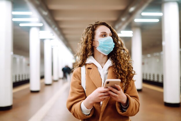 Chica en máscara médica protectora estéril con un teléfono en la estación de metro