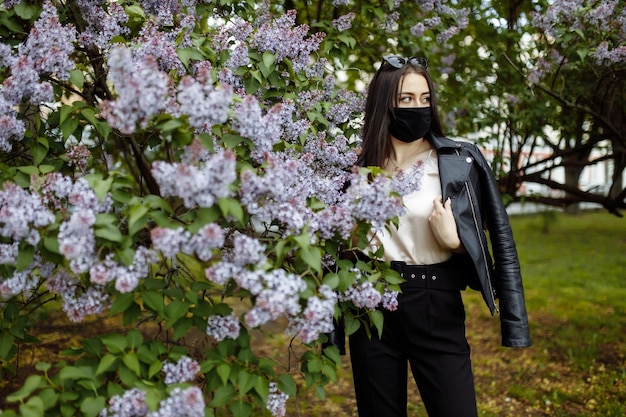 Chica en una máscara médica sobre un fondo de flores lilas protección de máscara negra contra el virus de la gripe co ...