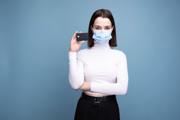 Chica en una máscara médica con una tarjeta bancaria en una pared azul.