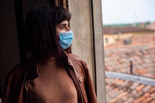 Foto chica con máscara médica en la ventana de su casa durante el período de cuarentena de covid