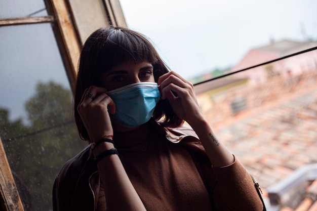 Chica con máscara médica en la ventana de su casa durante el período de cuarentena de covid