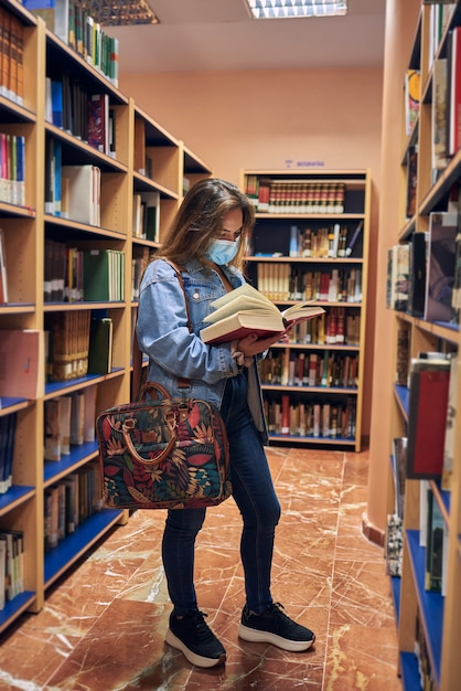 Foto chica con una máscara mira un libro entre los estantes de libros de una biblioteca