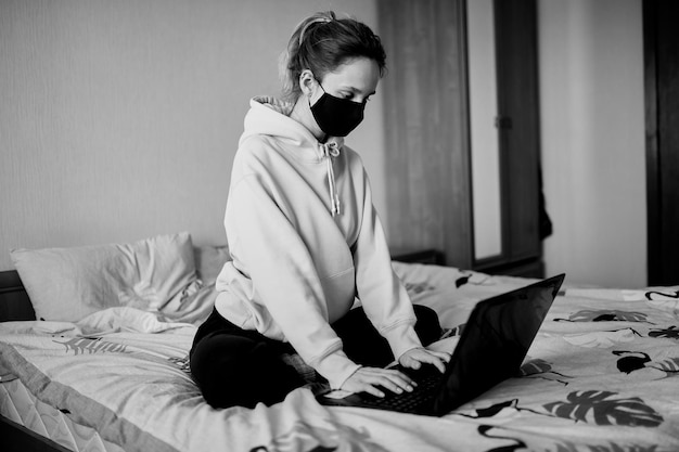 Chica con una máscara negra trabaja en una computadora portátil en casa aislada foto en blanco y negro