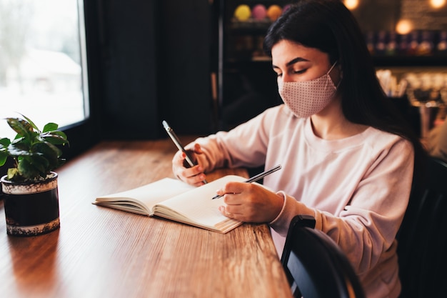 Foto chica en mascarilla, leyendo el teléfono y escribiendo en el cuaderno.