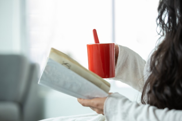 Chica mexicana bebiendo café en taza roja y leyendo un libro junto a la ventana