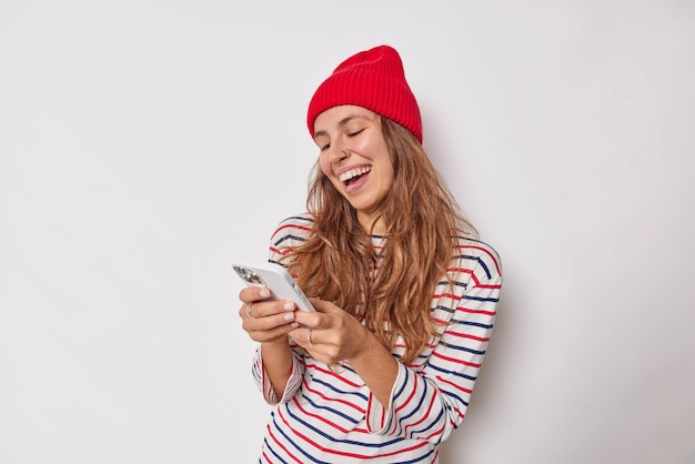 Chica milenaria alegre usa teléfono móvil moderno envía mensajes de texto chats en línea viste jersey de rayas de sombrero rojo tiene piercing en la nariz aislado en blanco. Concepto de personas y tecnología