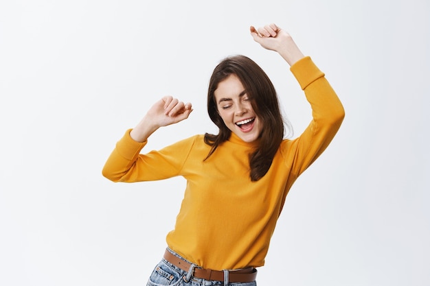 Foto chica milenaria llena de alegría bailando y divirtiéndose con los ojos cerrados y una sonrisa feliz, levantando las manos y relajándose al ritmo de la música, de pie contra la pared blanca