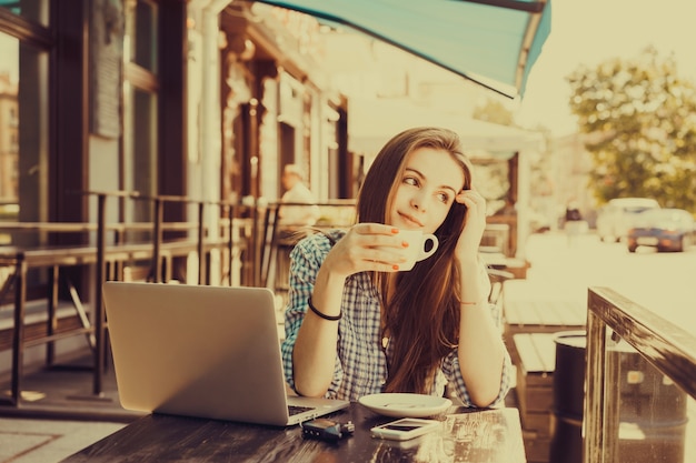 Chica mirando a la calle con una taza de café en la mano y un portátil delante